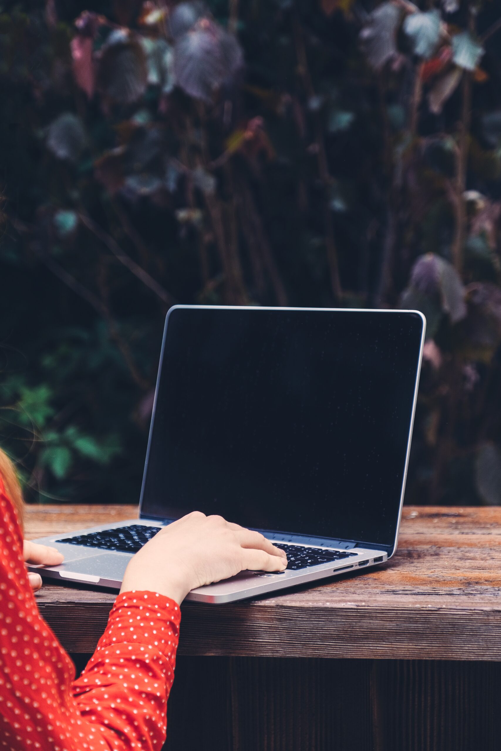 PERSON IN RED TOP TYPING ON LAPTOP AT WOODEN TABLE OUTSIDE