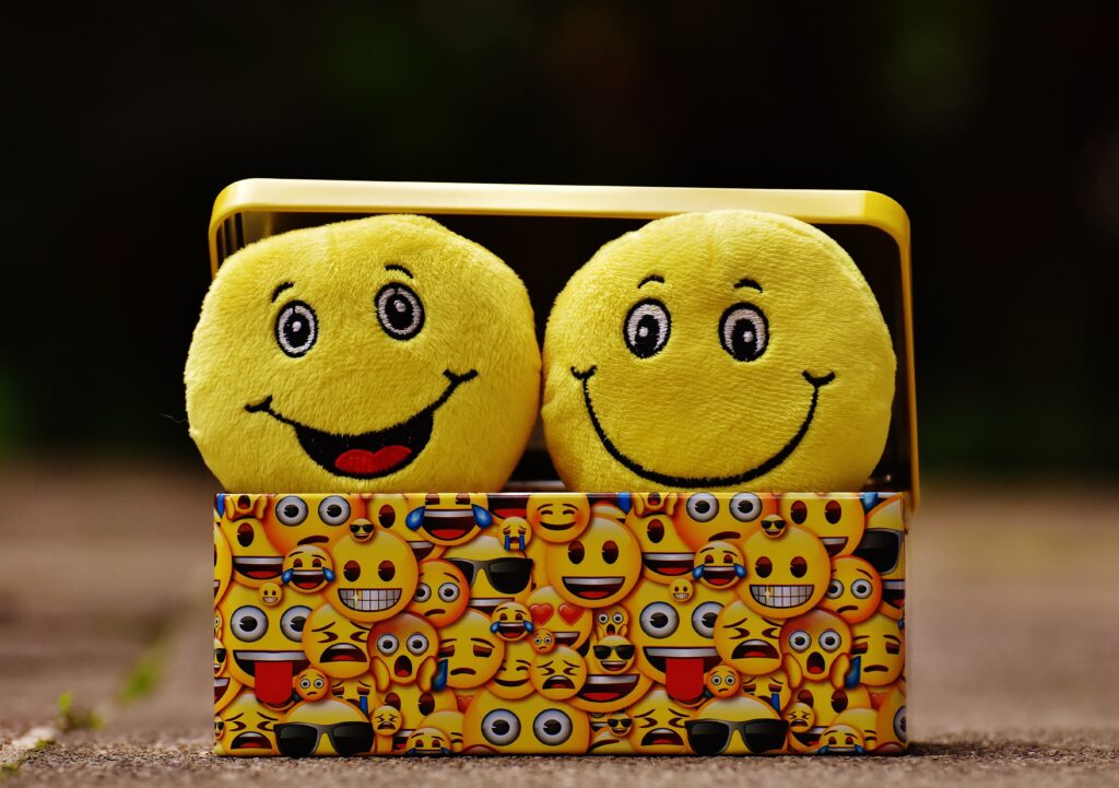 Two soft-toy smiling emoji cushions peeping out of a box covered with smiling emoji paper